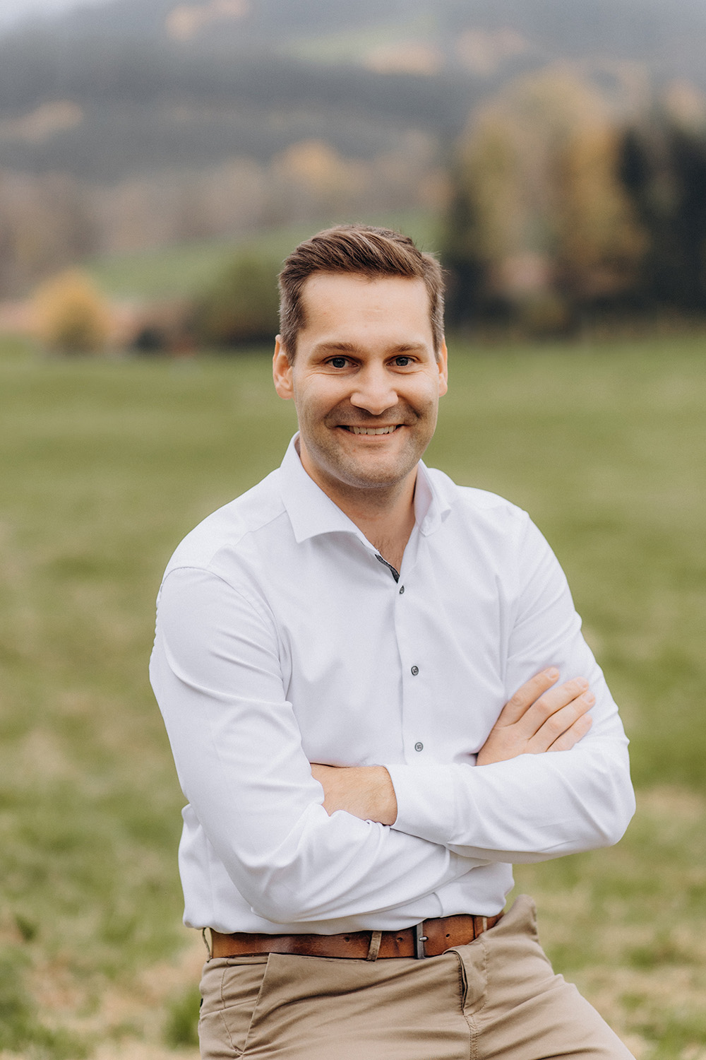 Ein Portrait von Raphael Wenzl. Er trägt ein weißes Hemd und Khakihosen. Er lächet auf einem offenen Feld. Im Hintergrund sind verschwommene Bäume und ein Hügel zu sehen, was auf eine ruhige, natürliche Umgebung schließen lässt.